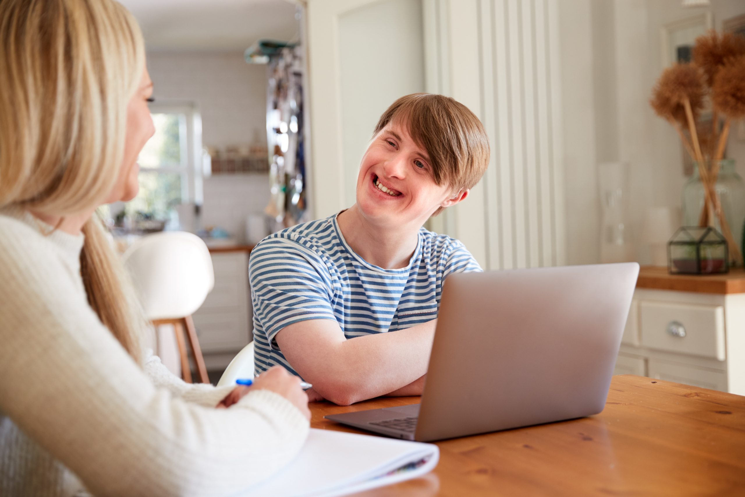 Man Who Has Downs Syndrome Sitting With Home Tutor Using Laptop For Lesson At Home